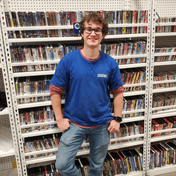 A male with curly brown hair wearing glasses is standing in front of a DVD stand wearing a blue "Goodwill" shirt.