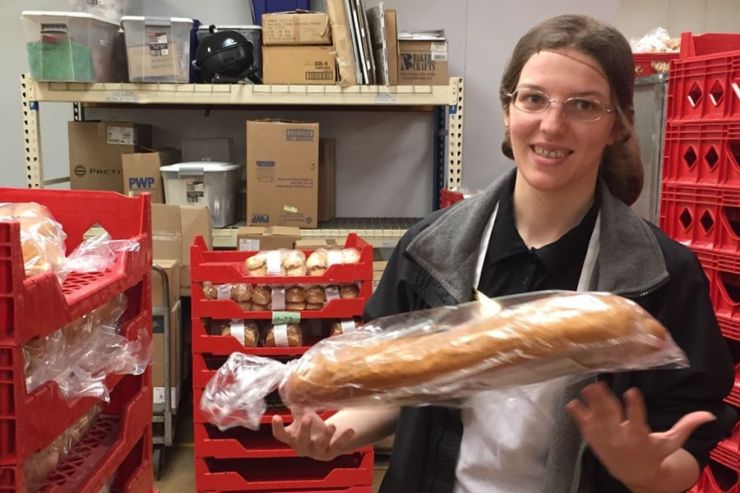 Photo of Cara smiling, wearing hairnet and tossing a wrapped baguette