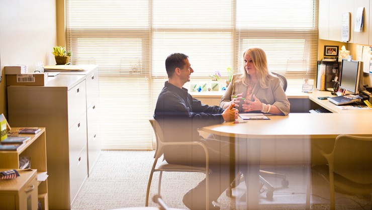 Photo of a man and woman sitting and talking at a desk in an office.