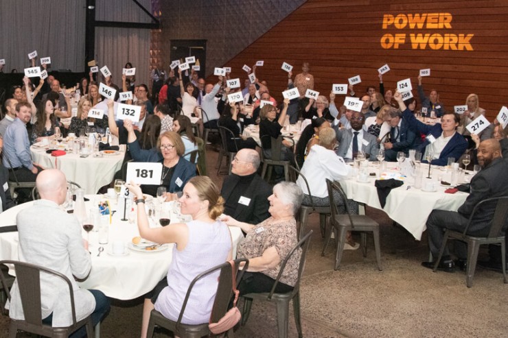 Photo of the multiple circular tables with people seated at them raising their cards to bid.